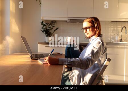 Frau Freiberuflerin in Brille mit Laptop und macht Notizen in einem Notizbuch, arbeitet während der Quarantäne wegen Coronavirus von zu Hause aus, hält Kaffee/Tee. Koz Stockfoto