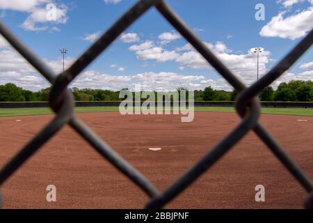 Blick über ein Baseballfeld von hinter der Heimatplatte durch eine der rautenförmigen Öffnungen in einem Kettengliedzaun an einem sonnigen Frühlings-Nachmittag Stockfoto