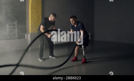 Junger Mann mit Ausbilder, der Kampfseile im Fitnessstudio verwendet Stockfoto