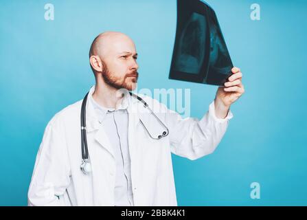 Attraktiver freundlicher kahler bärtiger Arzt, der die auf blauem Hintergrund isolierte Röntgenaufnahme betrachtet Stockfoto
