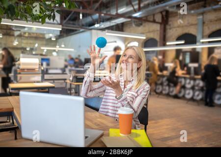 Blonde Frau sitzt am Laptop und spielt mit kleinem Ball Stockfoto