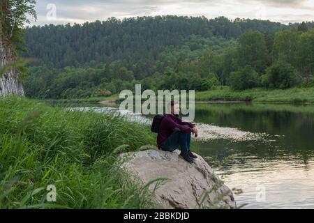 Der Touristenmann genießt einen schönen Blick auf die Natur von Wald, Fluss und Bergen. Junger bärtiger Kerl, der auf einem großen Stein am Ufer des Gebirgsflusses sitzt. Stockfoto