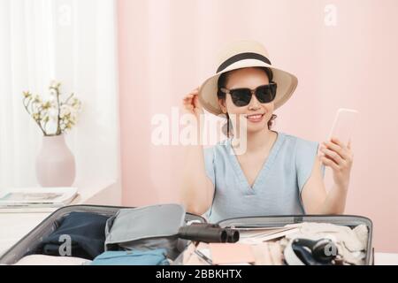 Junge Frau, die sich für einen Hut entscheidet, der mit Kleidung und Koffer auf dem Bett sitzt. Vorbereitung für den Sommerurlaub Stockfoto