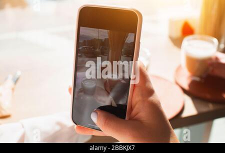Junge Frau nimmt ein Foto am Telefon auf. Weibliches Modell fotografiert eine Tasse Kaffee aus nächster Nähe Stockfoto