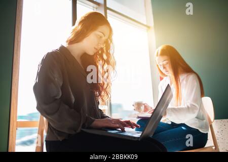 Weibliche Modelle arbeiten in einem Café. Eine junge Frau liest gerade ein Buch und ihre Freundin arbeitet auf dem Laptop. Stockfoto