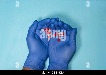 Die Hände des Arztes in blauen medizinischen Handschuhen sind nach vorne gestreckt und halten eine rote und weiße Tablette. Stockfoto