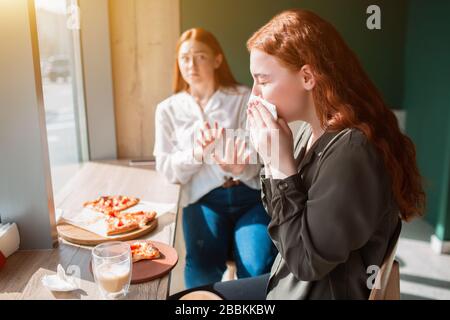 Junge Frau bläst in einem Café die Nase. Weibliches Modell sitzt am Tisch und wischt ihre Nase Stockfoto