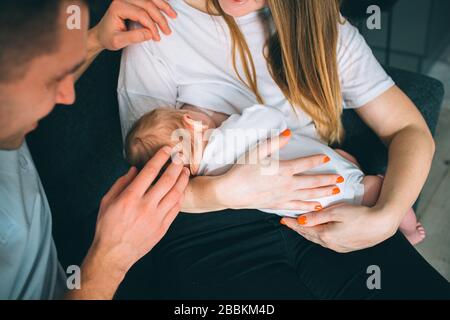 Vater und Mutter in einem Wohnzimmer mit einem neugeborenen Baby. Das erste Lebensjahr. Pflege und Gesundheit. Mutter hält ein kleines Kind, und Vater sitzt Stockfoto