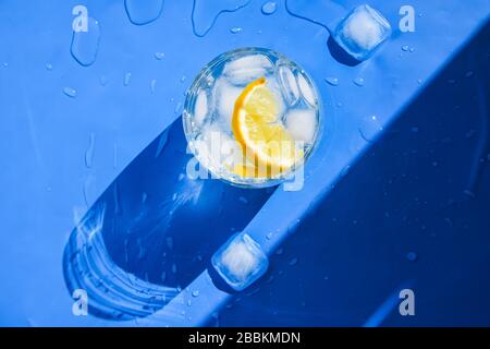 Ein Glas erfrischendes Wasser mit Eis und Zitrone auf blauem Grund. Wärmekonzept, frisch. Natürliches Licht. Flaches Layout, Draufsicht, Kopierbereich Stockfoto