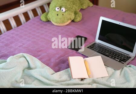 Computerlaptop mit Notizblock, Stift, Handy und weichem Spielzeug auf dem Bett im Schlafzimmer. Arbeiten Sie zu Hause. Stockfoto