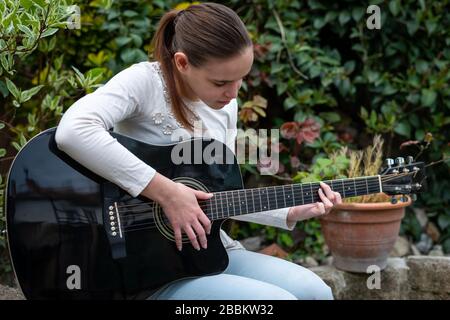 Natürlich aussehendes Teenager-Mädchen, das Gitarre im Garten spielt, lernt zu spielen, Hobby. Stockfoto