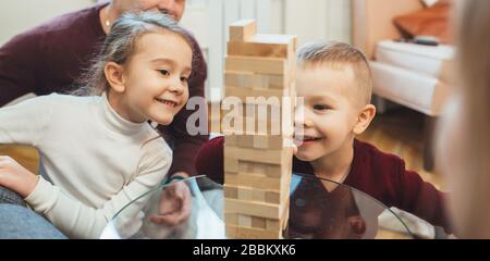 Zwei kaukasische Geschwister spielen zusammen jenga, während ihr Vater sie beobachtet Stockfoto