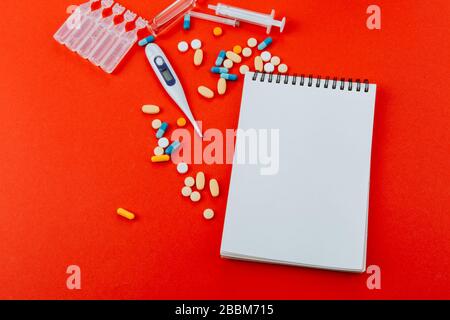 Notepad, eine Vielzahl von Medikamenten, Thermometer, Spritze und Ampullen mit Medikamenten auf rotem Hintergrund. Stockfoto