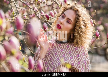 Porträt eines blonden schönen Mädchens mit lockig langen Haaren. Frau spaziert im Garten der blühenden rosafarbenen magnolie Stockfoto