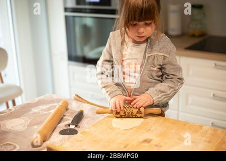 3 Jahre altes Kindermädchen in weißer Küche, das den Pizzateig mit einem Rollstift auf einem Holzbrett abflacht. Idee zu Lockdown-Aktivitäten. Stockfoto