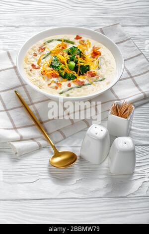 Portion Sahnesuppe mit Brokkoli, grüner Bohne, gebratenem Speck und geschreddeltem Cheddar-Käse in einer weißen Schüssel auf einem Holztisch mit goldenem Löffel, vertica Stockfoto