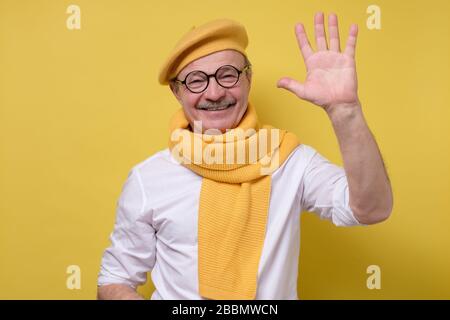 Älterer Mann mit Brille und gelbem Hut und Schal, der hallo sagte und mit der Hand winkte. Stockfoto