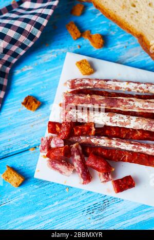 Hohe Winkelansicht einiger Fuet- und Chorizo-Stücke, zweier verschiedener spanischer Wurstwaren, auf einer weißen Platte auf einem blauen Holztisch neben einigen Stockfoto