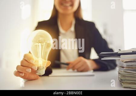 Eine Frau in einem Business-Anzug hält eine leuchtende Lampe in den Händen an einem Tisch im Büro. Stockfoto