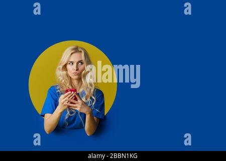 Porträt der jungen überraschten Frau, die über einem blauen Hintergrund steht. Mädchen pep aus einem Loch in der Wand. Stockfoto