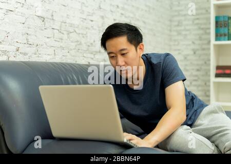 Junger, lächelnder asiatischer Mann in legerer Kleidung auf dem Sofa, der auf einem Laptop-Computer arbeitet. Freiberuflicher männlicher Mitarbeiter, der von zu Hause aus am Notebook arbeitet. Heimarbeit Stockfoto