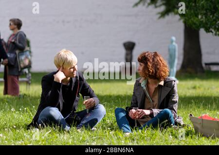 Zwei junge Frauen, die auf einem Gras sitzen, Weingläser mit Champagner halten und in einem Stadtpark kommunizieren Stockfoto