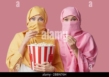 Zwei junge muslimische araberinnen tragen bunte Hijabs vor pinkfarbenem Hintergrund. Popcorn im Kino zu essen Stockfoto