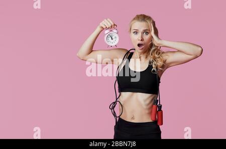 Zeit für Fitness. Schockierter Trainer der jungen Sportfrau hält pinkfarbenen Wecker in der Hand Stockfoto