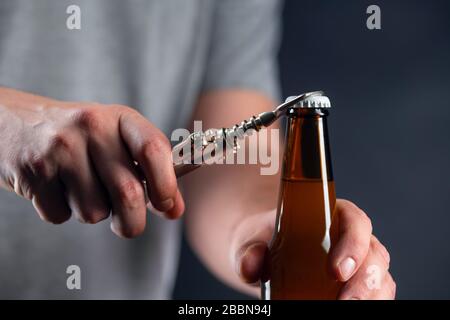 Männer öffnen kalte Bierflasche mit Kappe auf schwarzem Hintergrund. Hände knacken gekühlten Weizen oder Lagerbier mit einem Öffner auf dunklem Hintergrund Stockfoto