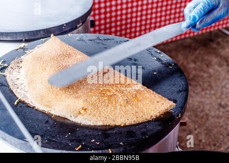 Crepes Pfannkuchen auf einem Lebensmittelmarkt machen. Eine Hand macht herzhafte Krepes im Freien auf einem Metallgrusel. Stockfoto