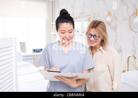 Asiatische Kosmetikerin in Schrubben füllt Informationen über den Kunden aus, während sie vor dem kosmetischen Verfahren mit einer reifen Frau spricht Stockfoto