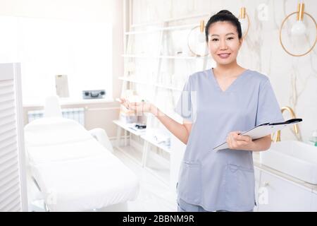 Portrait des lächelnden asiatischen Kosmetologen in den mit Zwischenablage stehenden Schruben und lädt Sie ein, Hautpflegeverfahren durchzuführen Stockfoto