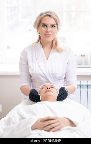 Portrait junger Kosmetologe in schwarzen Handschuhen mit Wattepad beim Reinigen der Gesichtshaut reifer Frauen vor dem Eingriff Stockfoto