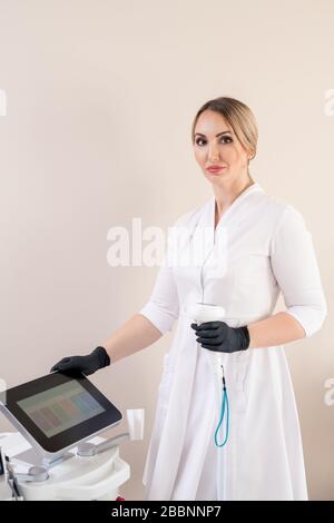 Porträt einer ernsthaften Kosmetikerin im Labormantel und chirurgische Handschuhe, die mit Griff an der Laser-Haarentfernungseinrichtung stehen Stockfoto