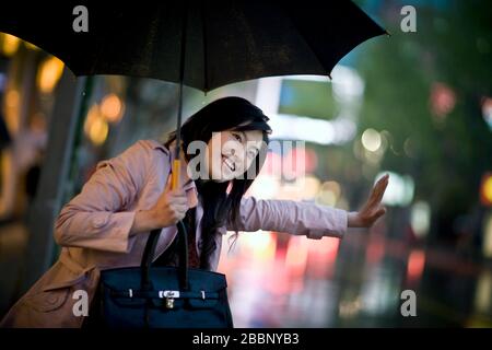 Junge Erwachsene Geschäftsfrau, die ein Taxi haelt, während sie einen Regenschirm hält. Stockfoto