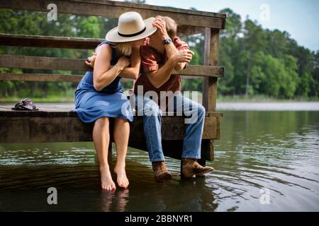 Reifer Mann lehnt sich romantisch an, um seine Frau zu küssen, während sie zusammen auf einem Holzsteg über einem See sitzen. Stockfoto