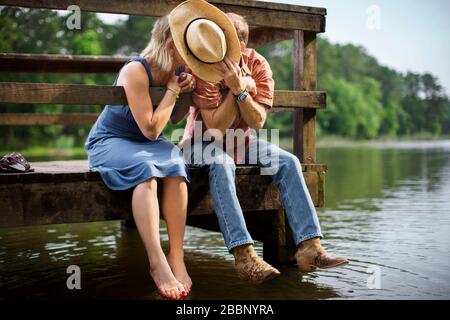 Reifer Mann hält einen Cowboyhut auf, um den Blick zu versperren, während er sich einlehnt, um seiner Frau einen romantischen Kuss zu geben, während sie zusammen auf einem Holzsteg über einem See sitzen. Stockfoto
