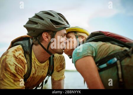 Schlammige junge Paare teilen sich einen Kuss, während sie eine Pause vom Mountainbiking an einem See machen. Stockfoto