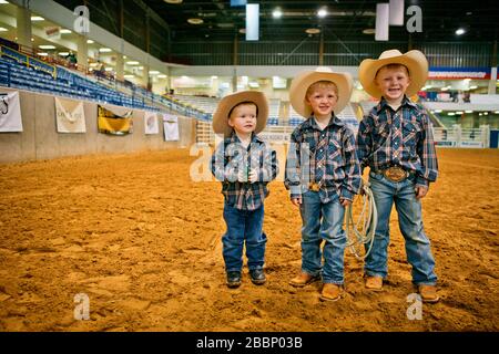 Porträt von drei lächelnden jungen Jungen, die als Cowboys in einer Rodeo-Arena gekleidet sind. Stockfoto