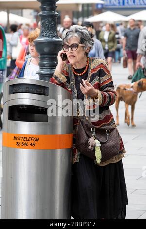 Eine attraktive, gut gekleidete ältere Frau hat ein animiertes Handy-Gespräch auf einer überfüllten Straße in Wien, Österreich. Stockfoto