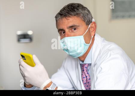 Selbstbewusster Arzt, der in Coronavirus-Zeiten Maske und Handschuhe im Medizinstudio trägt und sein Smartphone putzt. Stockfoto
