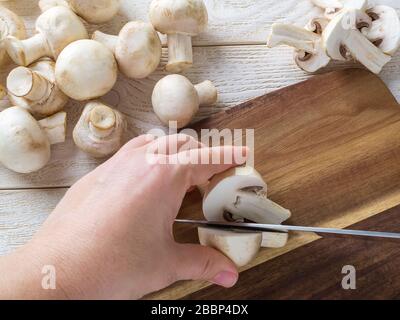 Frau hält einen Champignon halbiert mit scharfem Messer auf einem Holzschneidbrett. Pilze als pflanzliches Eiweiß, rohe Ernährung, Vegetarismus. Stockfoto