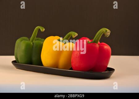 Drei Paprika in Rot, Gelb und Grün, deren Stiele in einer Keramikschale auf einem hellen Tisch und dunklem Hintergrund aufgereiht sind Stockfoto