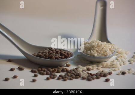 Zwei keramische Löffel mit Linsen und weißem Reis auf einer neutralen weißen Oberfläche, auf der Linsen und Reiskörner gemischt werden Stockfoto