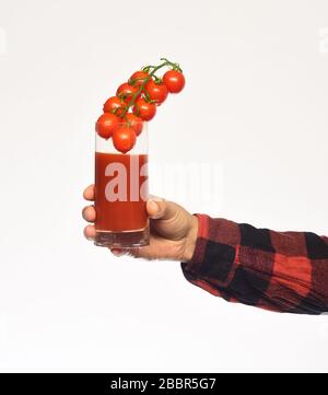 Das Handgelenk von Mans zeigt Tomaten aus roter Kirsche und ihr Produkt. Guy hält selbst erwachsene Ernte. Männliche Hand hält Glas Tomatensaft mit Beeren innen isoliert auf weißem Grund. Landwirtschaft und Herbstkonzept Stockfoto