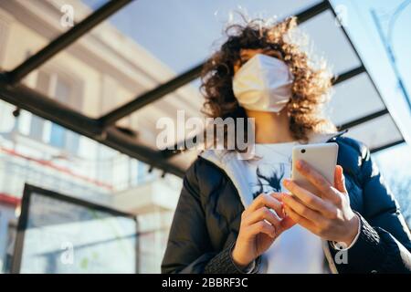 Junge Frau aus nächster Nähe, die eine schützende Gesichtsmaske trägt, die an öffentlichen Verkehrshaltestellen in der europäischen Stadt steht und ihr Smartphone hält. Stockfoto