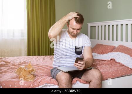 Wütender junger Mann, der von zu Hause aus im Schlafzimmer arbeitet, sein Telefon in der Hand hält und eine andere Hand in seinem Kopf steht Orange CAT neben ihm Stockfoto