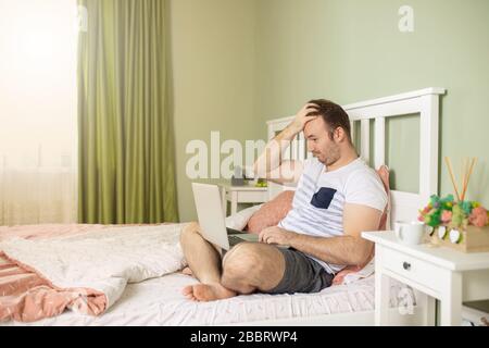 Der Mann mit einem Laptop im Bett erhielt schlechte Nachrichten, er ist verwirrt, hält die Hand am Kopf und beobachtet den Bildschirm in schockierten Augen. Stockfoto