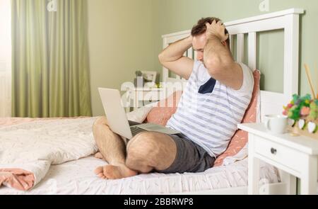 Der Mann mit einem Laptop im Bett erhielt schlechte Nachrichten, er ist verwirrt, hält die Hand am Kopf und beobachtet den Bildschirm in schockierten Augen. Stockfoto