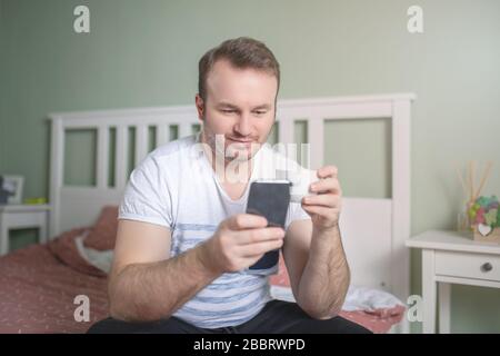 Fröhlicher Mann am Morgen auf dem Bett sitzend, während er Kaffee mit seinem Telefon in der Hand trinkt. Stockfoto